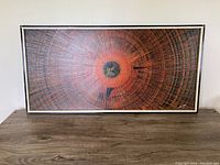 Large rectangular abstract oil painting on board resting on a wooden surface, showing concentric circular patterns in black, orange, and brown tones with a black and white frame.