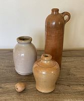 Photo showing three antique crocks on a wooden surface with a cork stopper in front, highlighting size and shape differences.