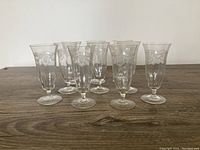 Set of ten antique etched crystal glasses arranged in a staggered formation on a wooden surface, showing clear etching with floral and vine motifs, flared rim and round base.
