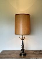 Front view of the lamp with the shade on, showing the tall cylindrical beige fabric shade and the turned brass base with twisted column detailing.