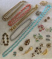 Top-down view of assortment of vintage costume jewelry including bead necklaces in turquoise, coral, gold-tone, and silver tones, assorted rhinestone brooches, earrings, and a black enamel bracelet arranged on gray surface.