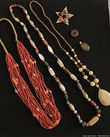 Overall view of all jewelry items on black background showing coral necklace, agate necklace, gold-tone necklace with pendant, star-shaped brooch, round brown bead, and oval pendant.