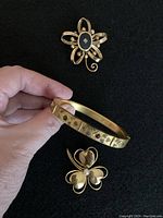 Hand holding an oval gold-tone bracelet inscribed Bates & B. with decorative hearts next to two gold-tone pins, one floral with black center and one heart-shaped with pearl