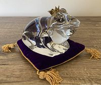 Front side view of the clear crystal frog paperweight sitting on the purple velvet cushion with gold tassels, showing details of the gold crown and crystal clarity.