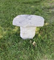 Outdoor solid carved stone pedestal seat seen from an angle, showing the rough carved base and smooth top seat surface on grass.