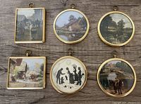 Top view of six brass framed miniatures arranged in two rows of three on wooden surface showing detailed painted scenes including cottages, village streets, rural landscapes and silhouette family scene.