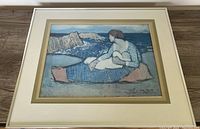 Framed artwork showing a mother holding a child by the sea, a vintage print with muted blue and earthy tones.