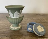 Photo showing the green Jasperware miniature urn-style vase and blue Jasperware lidded ring dish with classical white relief decorations on a wooden surface.