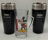 Photo shows one clear glass mug with Mickey Mouse design flanked by two black stainless steel Ford Taurus tumblers with silver rims and logos.