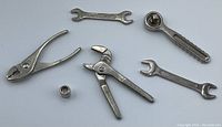 Five small silver-tone metal toy tools including toy pliers, pipe wrench, adjustable wrench, and two open-end wrenches plus a small metal nut.