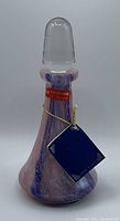 Clear photo of the Lavorazione T. Murano vintage perfume bottle showing its pink and blue swirled glass body, clear glass stopper, red label, and attached tag.