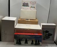 Photo showing multiple boxed miniature collectible village buildings and tree sets stacked together