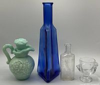 Photo shows four items: a tall cobalt blue glass bottle, a pale green Avon Skin So Soft carved container with lid, a small embossed medicine bottle, and a clear glass hen shot glass.