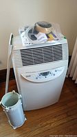 Front view of white DeLonghi PAC160 portable air conditioner with control panel and accessories in plastic bag on top, along with vent hose and adapter.