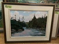 Framed oil painting showing landscape of a river flowing through trees and hills with cloudy sky