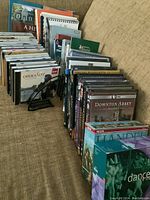 Wide view of mostly books, CDs and DVDs arranged on a couch or chair, showing lots of items packed close together in rows.