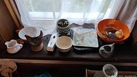 Overview of all items on a dark surface near a window: bowls, creamer, trivet, decorative bird bowl, plate, and orange bowl with items.
