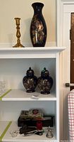 Photo of two matching black ceramic urns with lids on middle shelf, brass candlestick holder and tall black ceramic vase on top shelf, small items on lower shelf