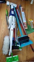 Various cleaning tools laid on wooden floor including Bissell Easy Vac, multiple brooms, dustpan, Swiffer mop, and duster.