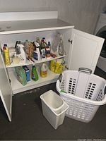 Wide view of white storage cabinet with cleaning liquids on shelves, white plastic laundry basket and two waste bins on floor