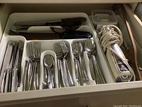 Top-down photo of flatware and cutting knives in organizer drawer showing knives, forks, spoons, and electric cutting utensil including cord and control switch.