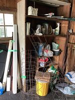 Photo showing several metal tomato cages, bags of soil and fertilizer, yellow plastic planter, garden spray bottle, and shelving with wicker basket and other supplies.