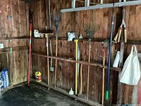 Garden tools hung on wooden wall showing pole trimmer, broom, various rakes, cultivator, pruners, and axe