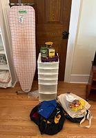 Front view of ironing board, plastic drawer unit with six drawers, decorative tins on top, backpack, shoulder bags, and grocery bags.