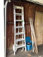 Full view of the aluminum ladder standing upright in a garage next to wooden sticks and plastic storage bins.