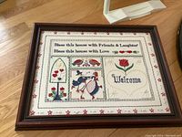 Framed needlepoint artwork with a welcome message, birds, flowers, and hearts, photographed on a wooden floor.