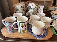 Set of multiple cups, mugs on a wooden cutting board with ceramic insert, showing various designs and handle styles.