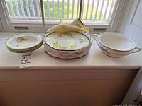 Set of Royal Doulton dishes including four bowls on left, six plates stacked in middle with fabric carrier, and serving dish without lid on right, displayed on window ledge.