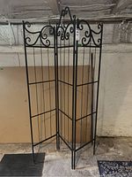 Three-panel black metal folding screen with decorative scroll pattern at top, grid lower design, folded in zig-zag shape in basement setting.