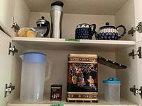 Top shelf showing French press without glass, insulated travel thermos, Polish blue and white ceramic teapot and creamer, and lemon juicer.