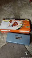 Boxed Black & Decker powered hand saw with metal toolbox closed in front on concrete floor.