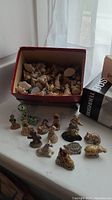 Collection of small ceramic and Wade Red Rose figurines including animals and characters shown arranged on a flat surface, with a storage box containing more figurines nearby.