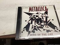 Front cover of the Metallica Hero Of The Day CD single showing jewel case and cover artwork with star-shaped black paint splatter design and band/title names.