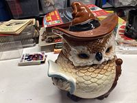 Front and side view of ceramic owl cookie jar with large blue eyes, detailed brown and cream feathers, wearing a large brown witch hat, holding a book.