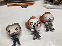 Three Funko Pop figures laid on table, one Joker and two Pennywise figures from It