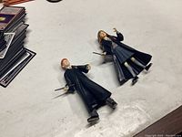 Two Harry Potter character figures, one with brown hair and one with red hair, both in black Hogwarts robes holding wands lying on a white surface.
