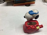 Ceramic Snoopy toothbrush holder main front and top view showing Snoopy head with blue cap and red base with embossed Snoopy text.