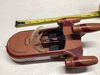 Brown and reddish plastic Landspeeder toy with clear canopy and two black seats shown from above with measuring tape for scale