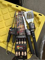 Photo of the new 7-piece BBQ tool set including tongs, fork, spatula, knife, and skewers displayed on a yellow surface.