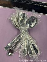 Photo shows two bundles of stainless steel utensils wrapped in clear plastic, one bundle of six forks and one bundle of six spoons.