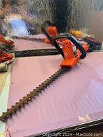 Full length view of the hedge trimmer showing orange body and long metal blade with teeth.