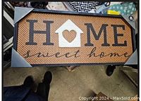 Photo shows a rectangular home decor wall sign with 'HOME' spelled out, a house shape replacing the letter 'O', and the phrase 'sweet home' below in script. The textured background appears woven, with metal corner accents.