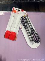 Photo of two silicone pastry brushes in red with white handles and two stainless steel whisks with black wires, all new and in packaging on a purple surface.