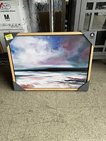 Photo of a medium-sized rectangular canvas print showing an ocean and sky scene with clouds in shades of blue, purple, pink, and white, framed in wood with protective corner covers.