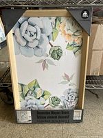Front view of the decorative magnet board with floral succulent design and wood frame, showing the packaging label with size and brand information.