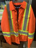 Front view of an orange high visibility work jacket with reflective stripes and snap button closure hanging on a blue hanger.